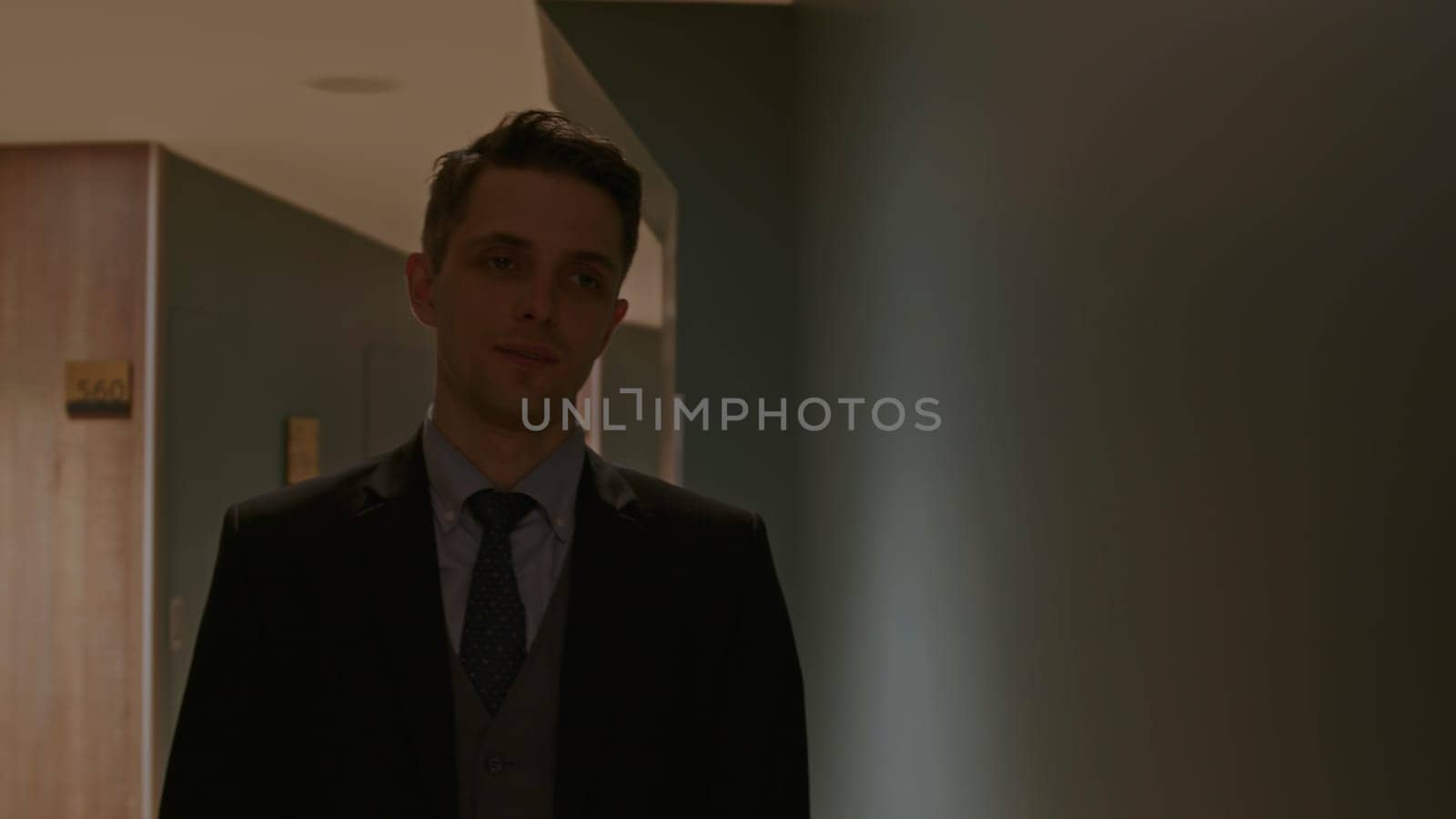 Security man walking along rooms in hotel corridor. Stock clip. Young man in suit in a dark building hall