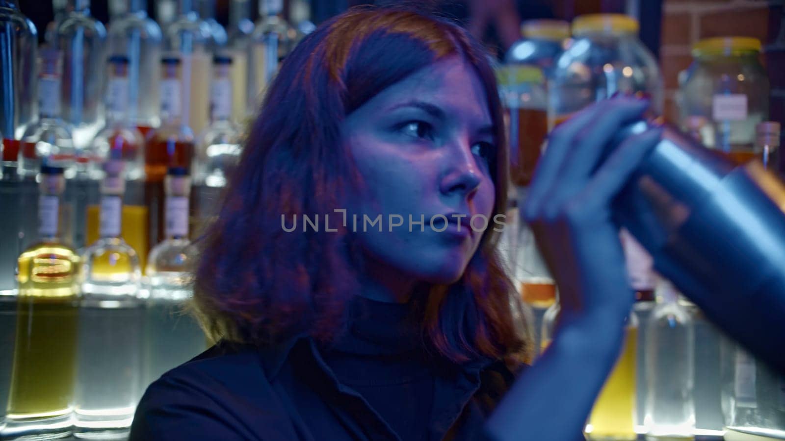 Young female bartender at nightclub. Media. Female bartender shaker drink at bar. Bartender prepares drink on background of many bottles of alcohol in bar.