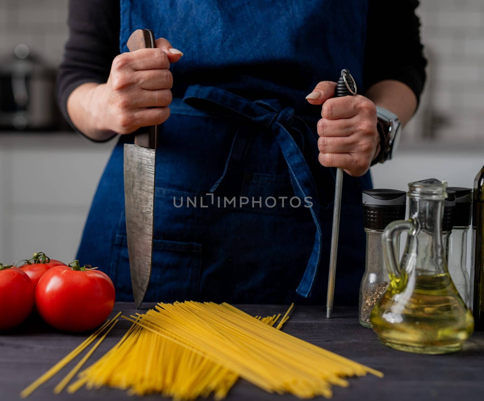 Large Kitchen Knife And Sharpener In Female Hands Are In Close-Up Over Table With Spaghetti, Tomatoes, Spices, And Basil