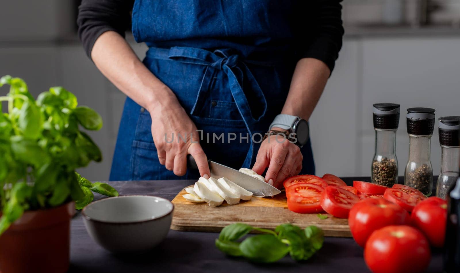 Preparing Caprese Salad, Female Hands Cut Mozzarella Cheese On A Board, With Mozzarella, Basil, And Spices Nearby