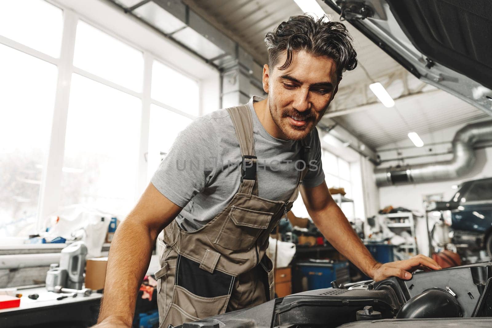 Young male mechanic examining engine under hood of car at the repair garage close up