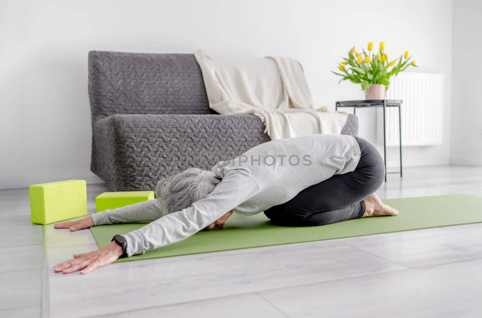 Grey-Haired Woman In baby Pose Practices Yoga At Home On The Floor In A Bright Living Room