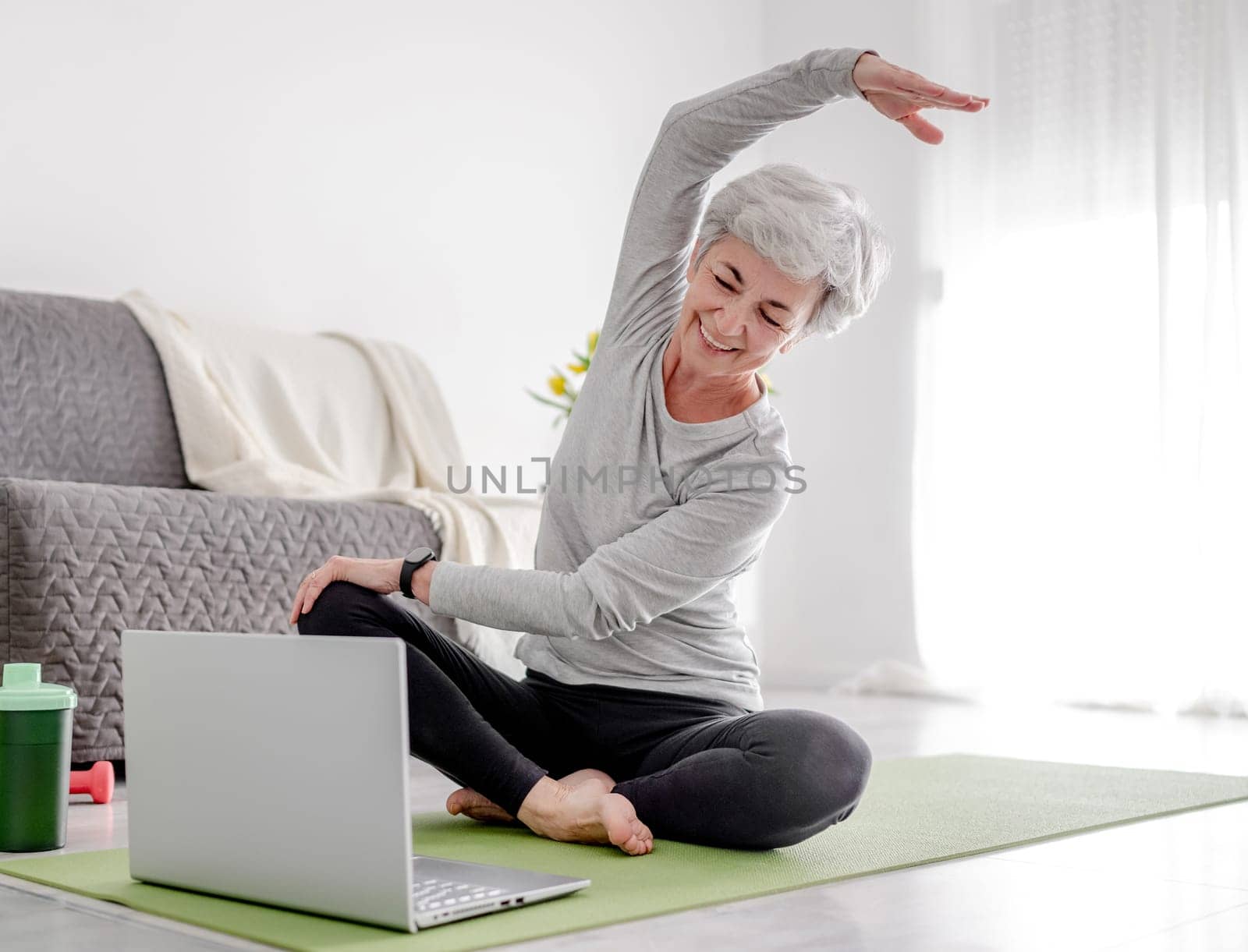 Pleasant 70-Year-Old Woman Exercises With Trainer Through Laptop, Doing Sports At Home On Yoga Mat