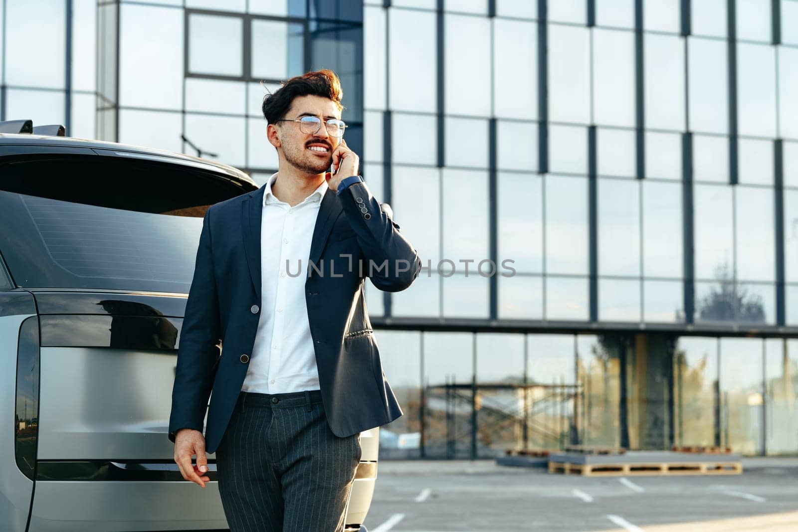 Young businessman in formal suit standing near luxury car and talking on the phone outdoors