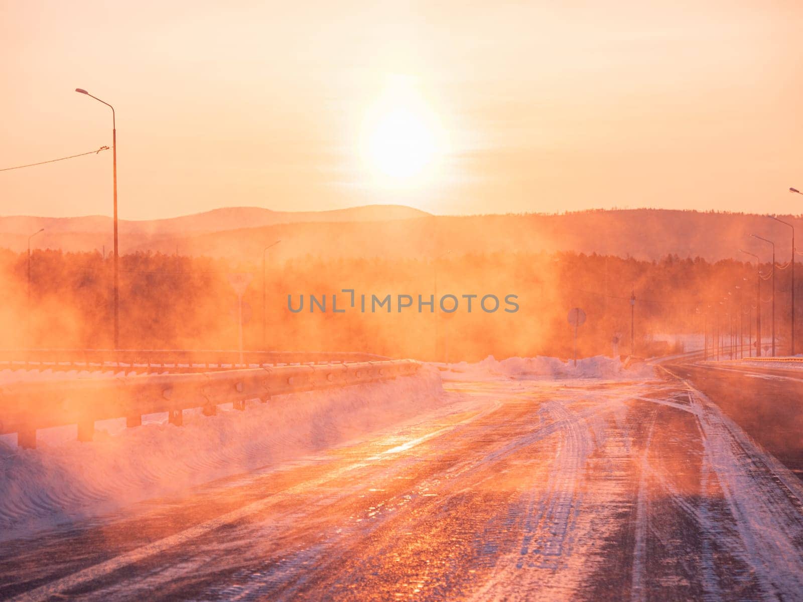 The sun rises beautifully over a snow-covered road in a serene winter countryside.