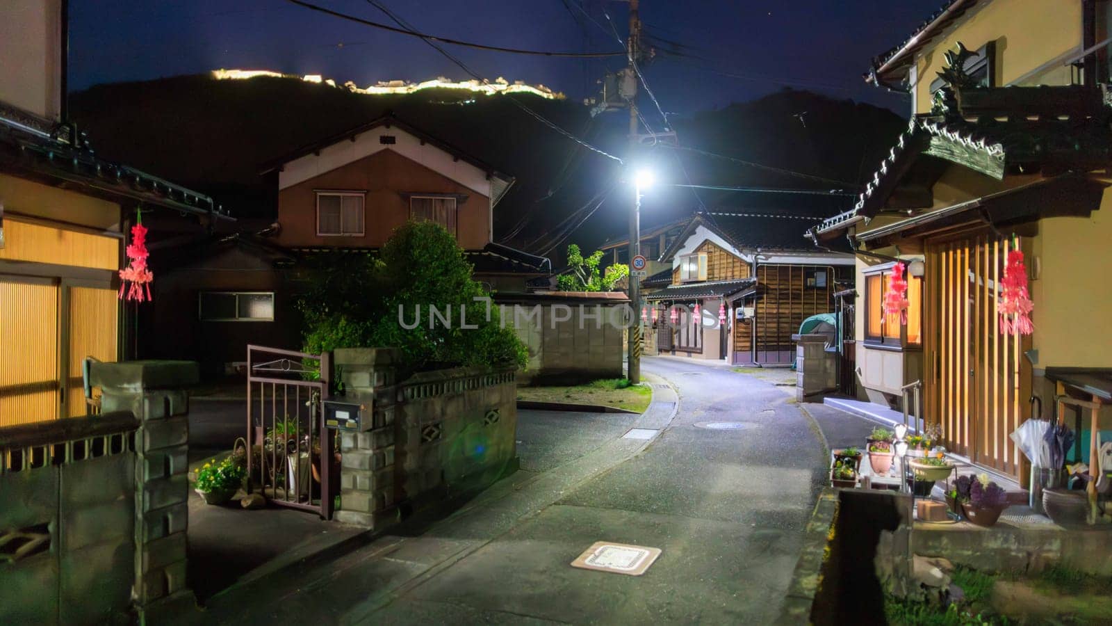 Traditional houses on empty road in Takeda at night with lit castle on hill by Osaze