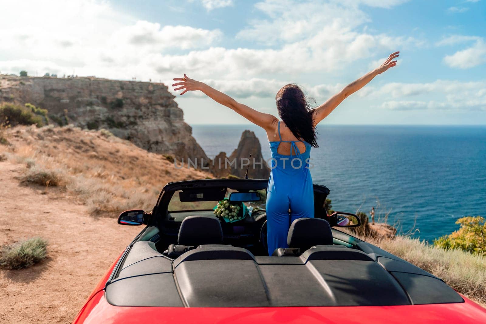 A woman is standing on the roof of a red convertible car, looking out at the ocean. She is smiling and she is enjoying the view