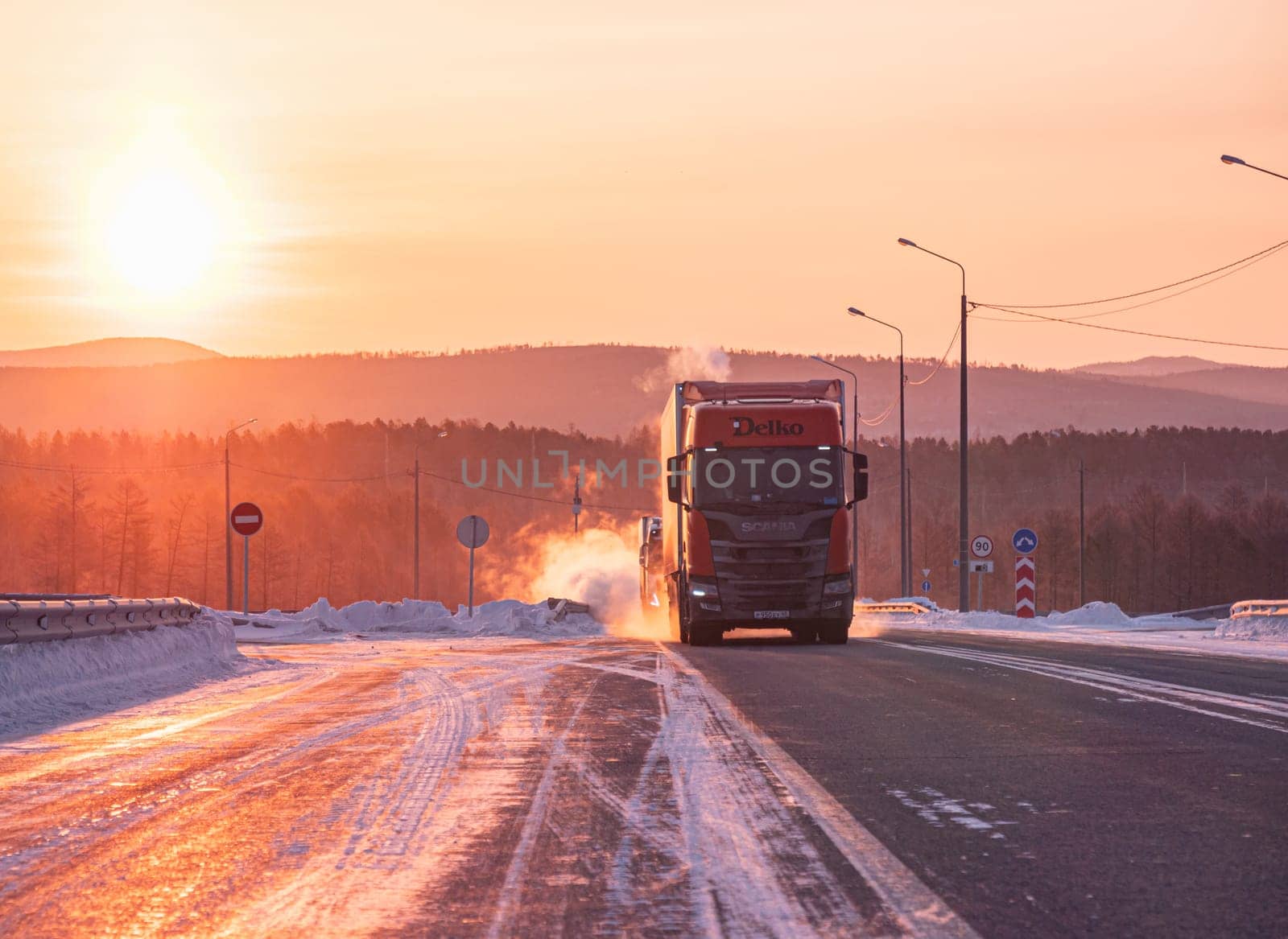 Two large trucks are driving along a snow-covered highway during a breathtaking winter sunrise.