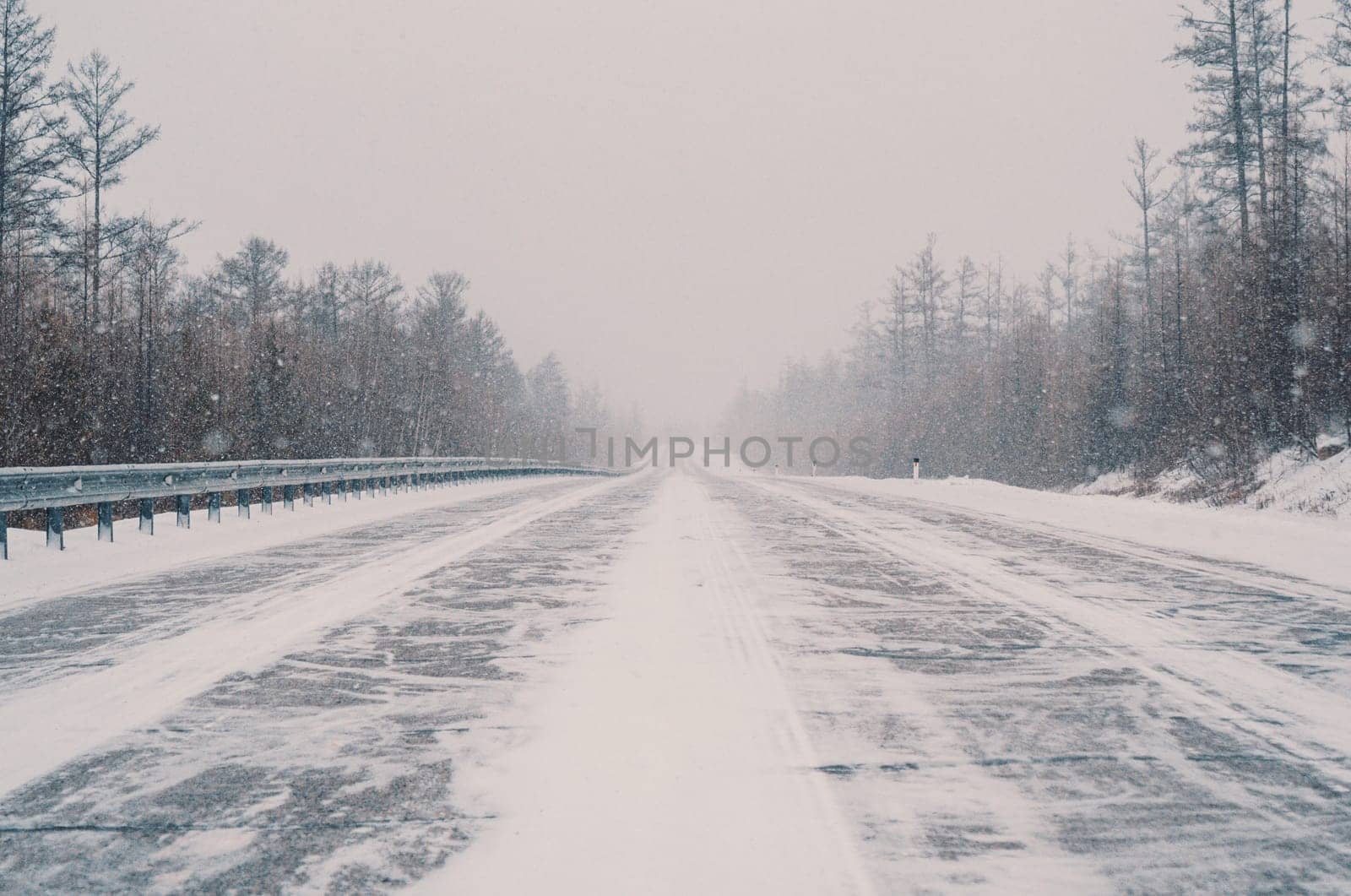 A remote highway runs through a dense forest, heavily covered in snow amid a fierce winter storm. Visibility is low, and the landscape is entirely white with streaks of snow crossing the road.