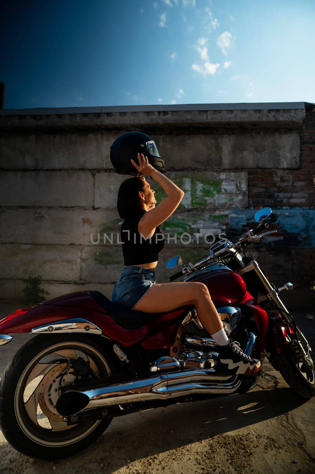 A brunette woman in denim shorts puts on a helmet while sitting on a red motorcycle. Vertical photo