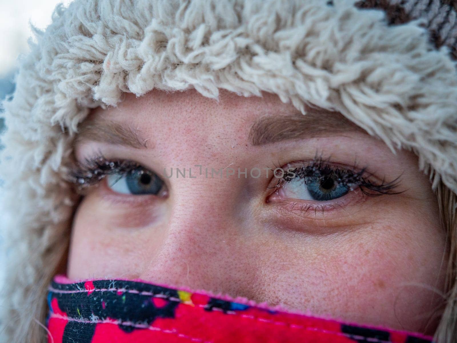 A close-up of a young womans face, showing her blue eyes and winter attire. She is bundled in a colorful scarf and a cozy knit hat, prepared for cold weather.