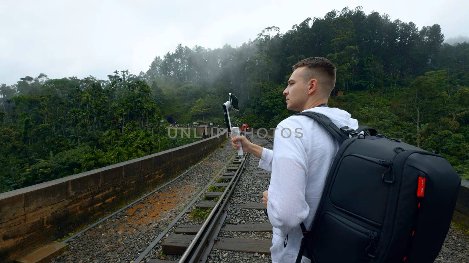 Rear view of a man tourist taking pictures of mountain forested landscape on smartphone. Action. Hiker walking on railways