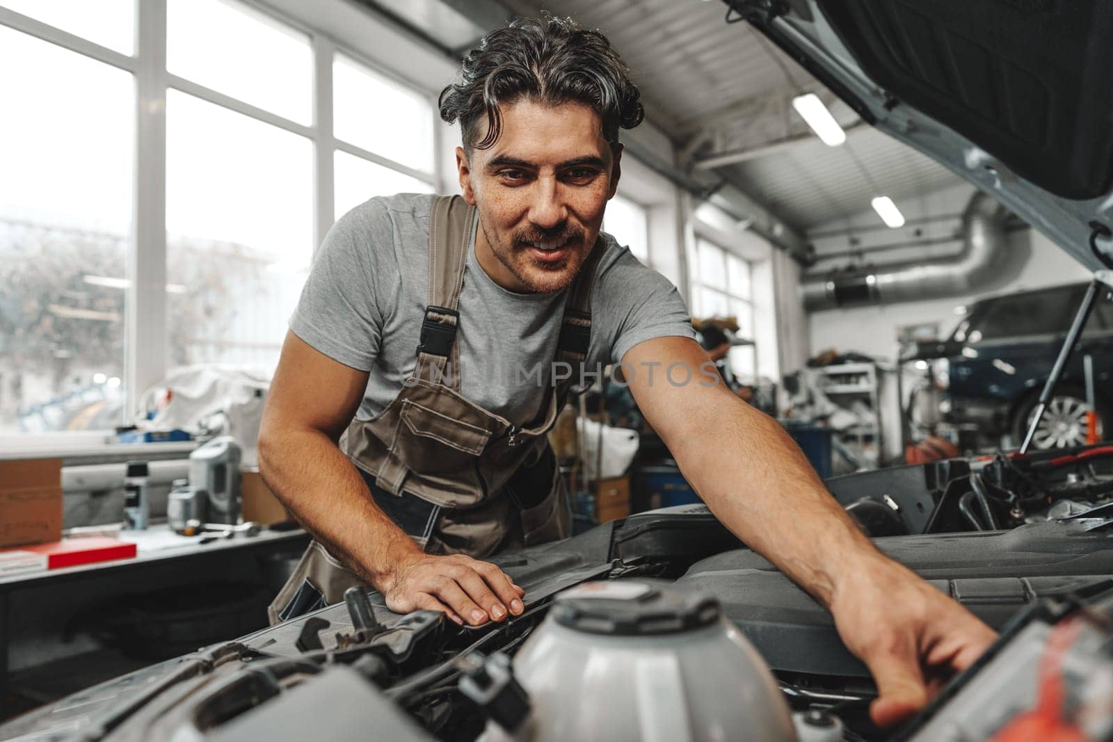 Young male mechanic examining engine under hood of car at the repair garage close up