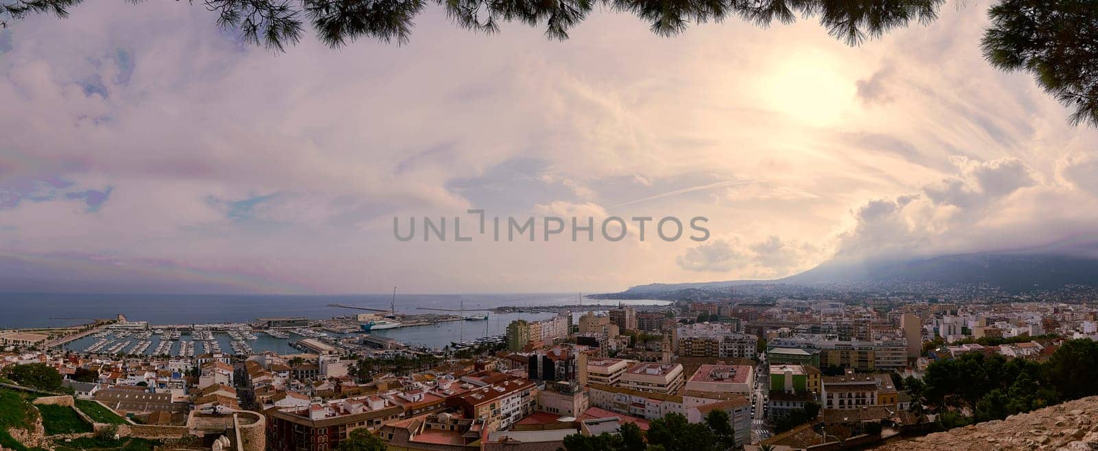 igh-resolution panoramic photograph showcasing the stunning coastal city of Denia, Spain, bathed in warm sunlight and adorned with fluffy clouds against a clear blue sky.