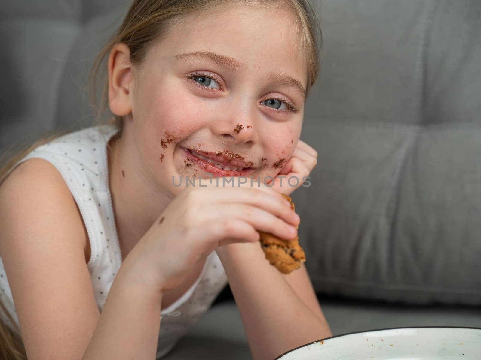 A cute little girl smeared in chocolate eats cookies while lying on the sofa