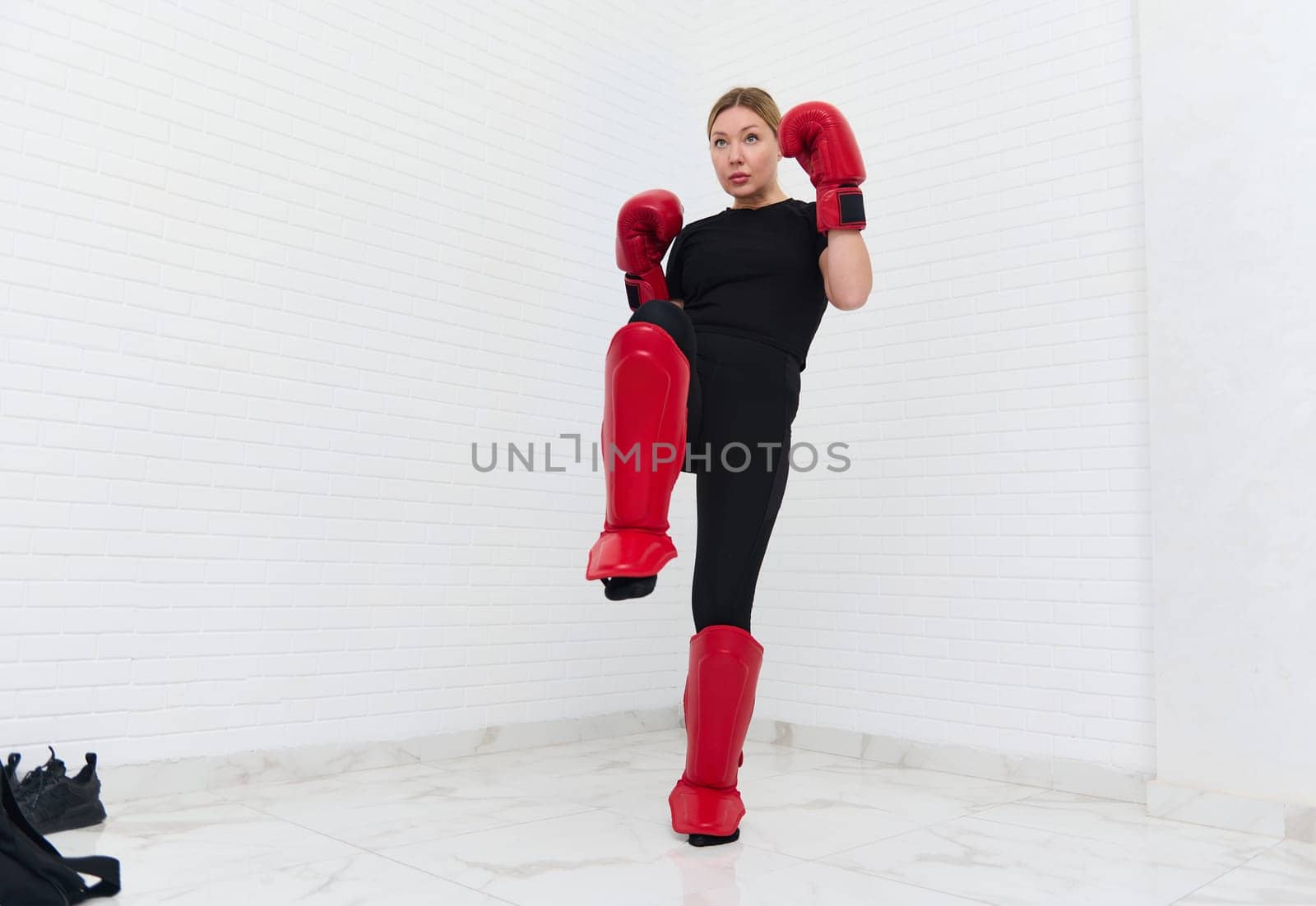 European blonde young kickboxing woman in black activewear and red kickboxing gloves, isolated on white background performing a martial arts kick. Sport exercise, fitness and cardio workout concept