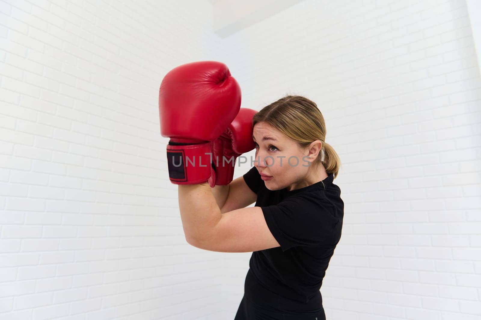 Portrait of a determined young female boxer in black sportswear and red boxing gloves, exercising, practicing box isolated over white background. Copy advertising space. People, sport and martial art
