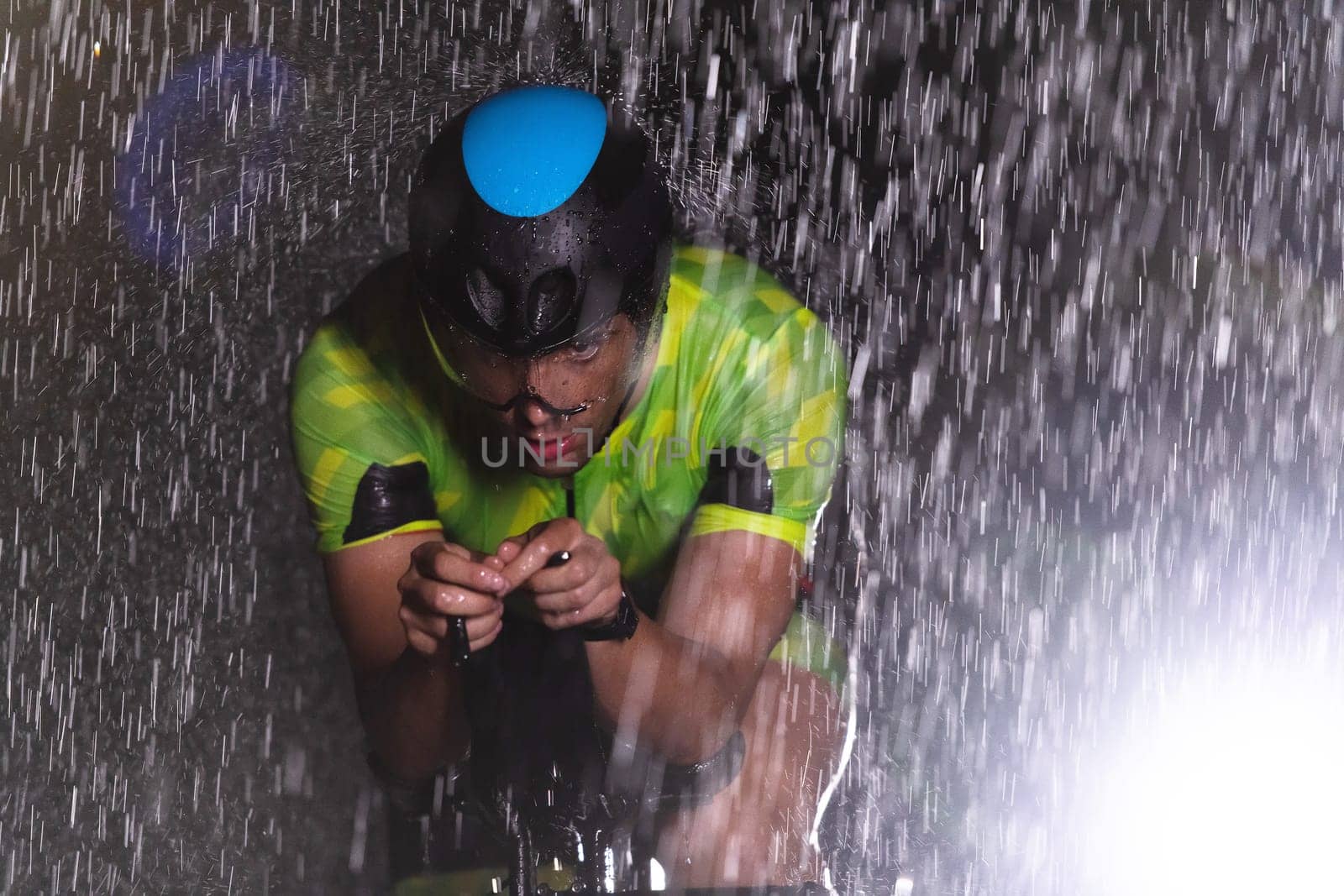 A triathlete braving the rain as he cycles through the night, preparing himself for the upcoming marathon. The blurred raindrops in the foreground and the dark, moody atmosphere in the background add to the sense of determination and grit shown by the athlete