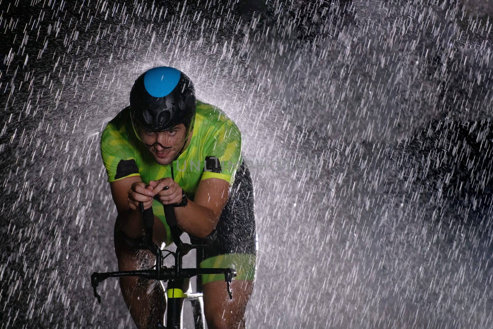 A triathlete braving the rain as he cycles through the night, preparing himself for the upcoming marathon. The blurred raindrops in the foreground and the dark, moody atmosphere in the background add to the sense of determination and grit shown by the athlete