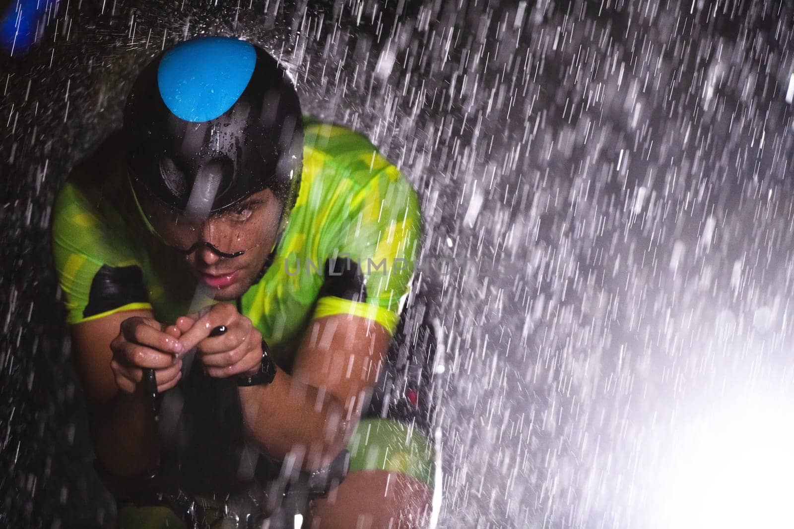 A triathlete braving the rain as he cycles through the night, preparing himself for the upcoming marathon. The blurred raindrops in the foreground and the dark, moody atmosphere in the background add to the sense of determination and grit shown by the athlete