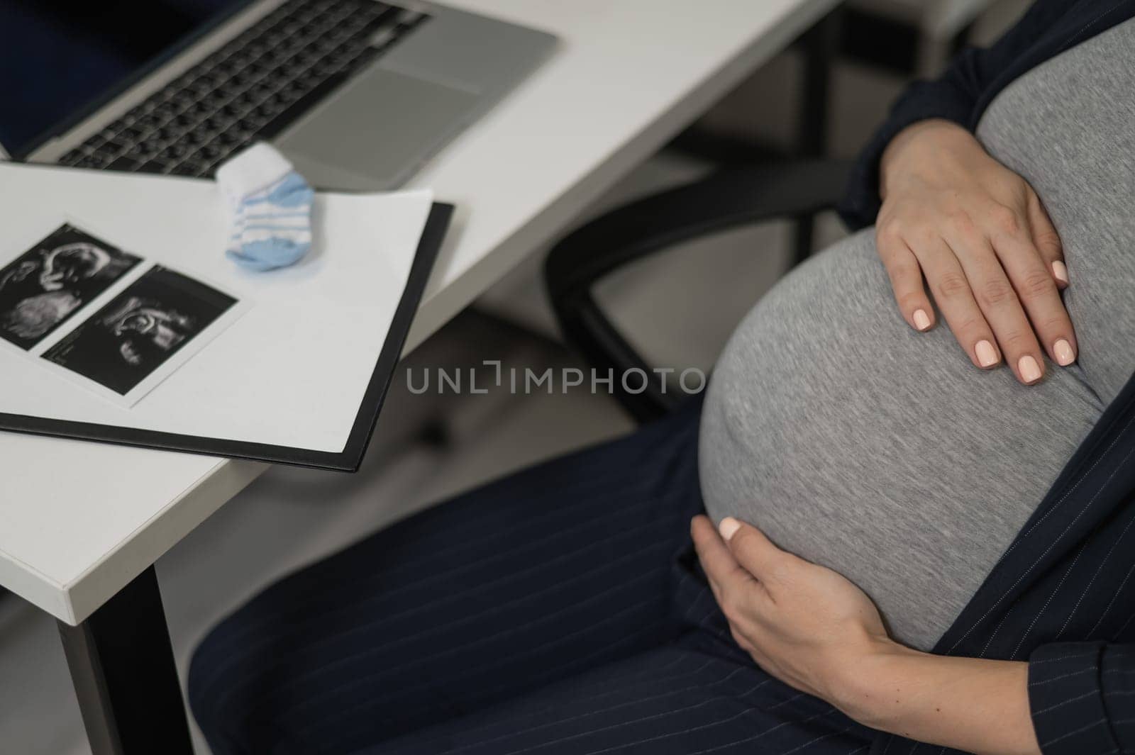 A pregnant woman works on a laptop in the office and looks at a photo from an ultrasound scan of the fetus. Belly close-up. by mrwed54