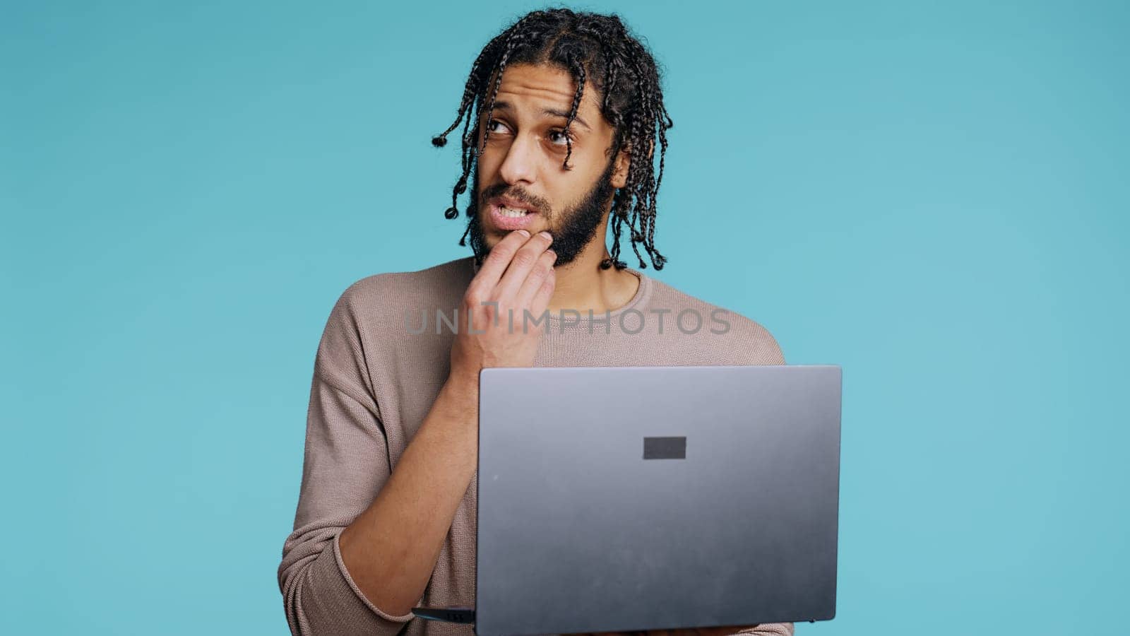 Middle Eastern man inputting data on laptop while standing in blue studio background. BIPOC employee typing on notebook keyboard, focused on solving project tasks, camera B