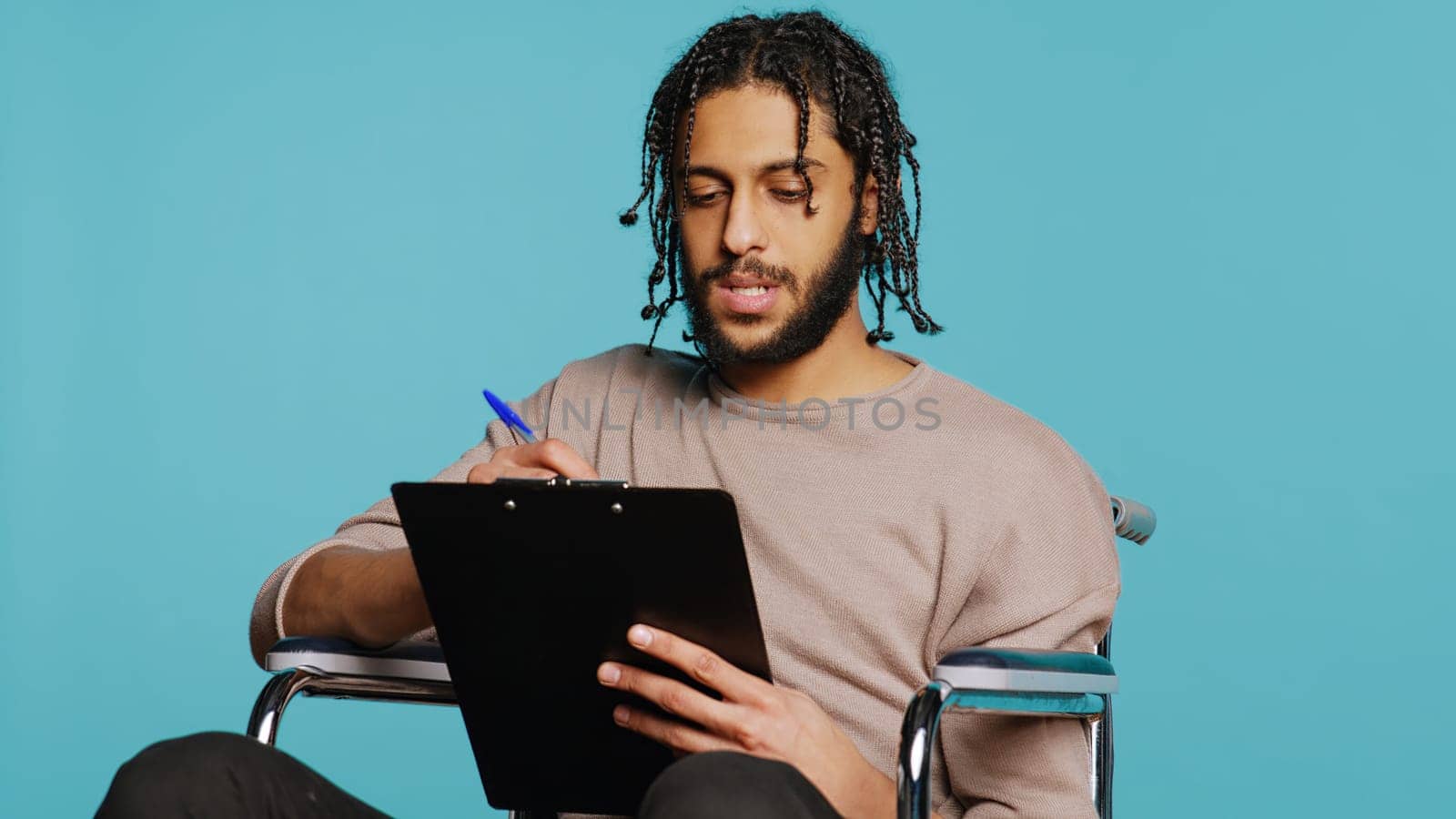 Middle Eastern man in wheelchair suffering from paraplegia holding clipboard in hand, using pen to write on paper. Person with disability completing forms, isolated over studio background, camera B