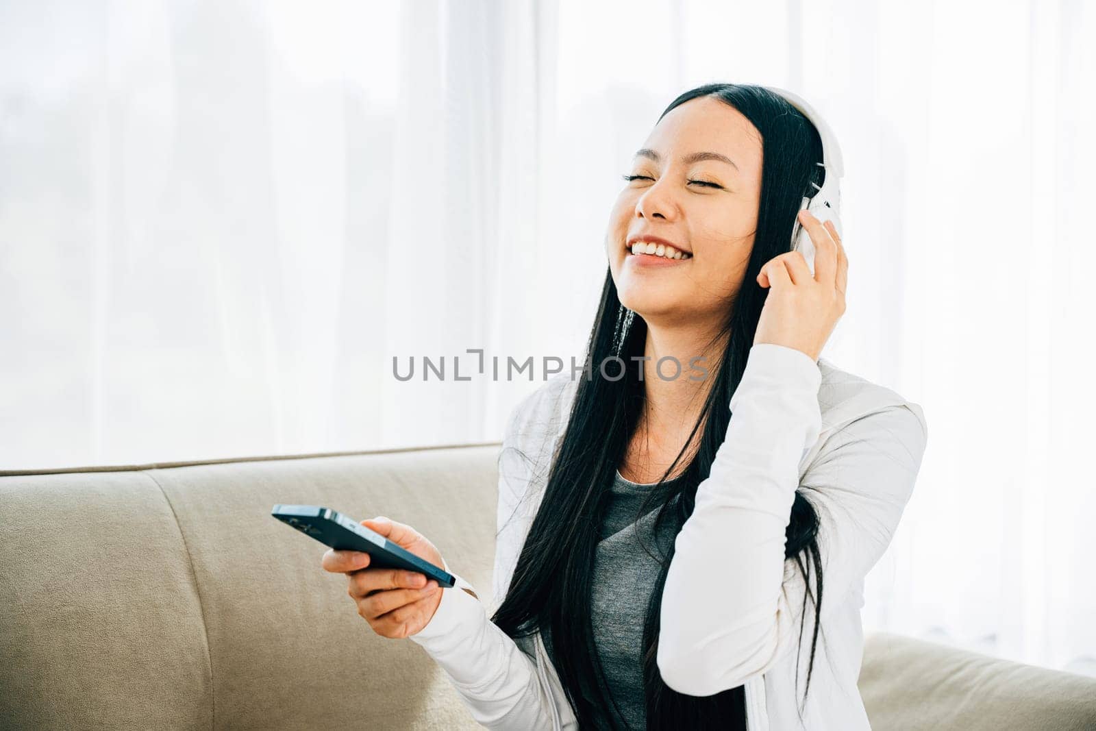 Attractive woman on sofa listens to music uses smartphone with headphones. Engaged in leisure relaxation and entertainment at home. Modern technology concept.