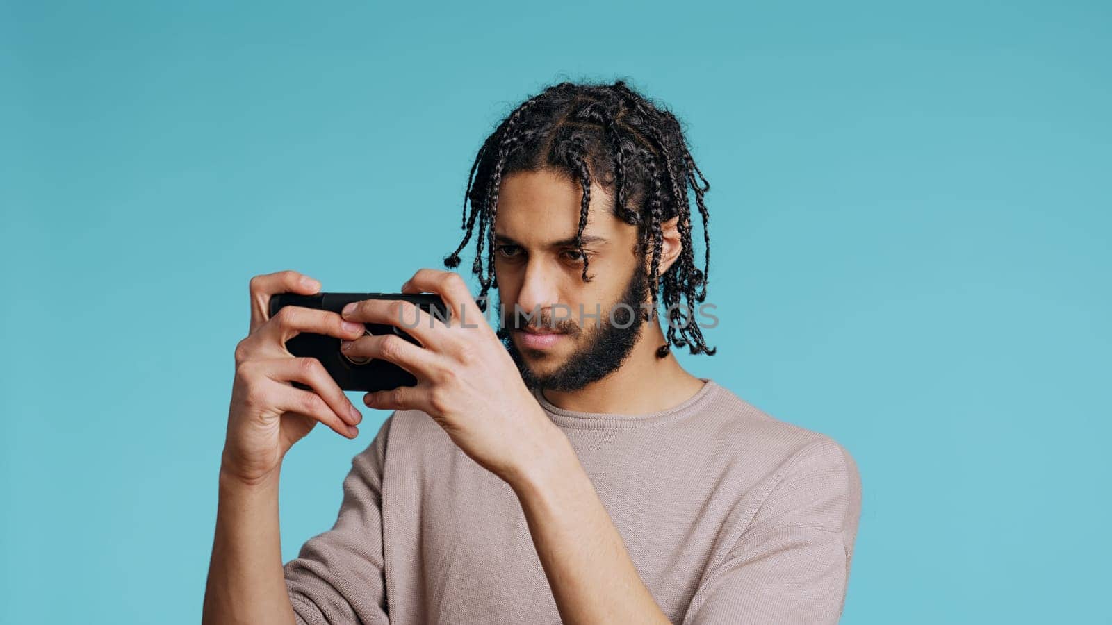 Gamer disappointed after receiving game over screen on cellphone touchscreen display. Middle Eastern man nodding head, upset after losing videogame, playing on phone, studio background, camera B