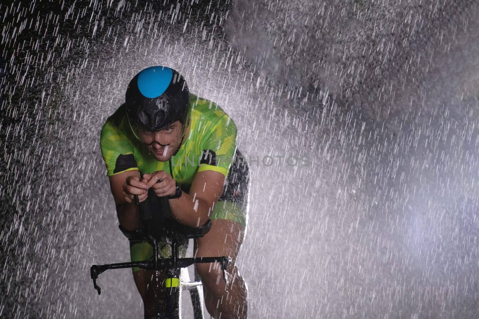 A triathlete braving the rain as he cycles through the night, preparing himself for the upcoming marathon. The blurred raindrops in the foreground and the dark, moody atmosphere in the background add to the sense of determination and grit shown by the athlete