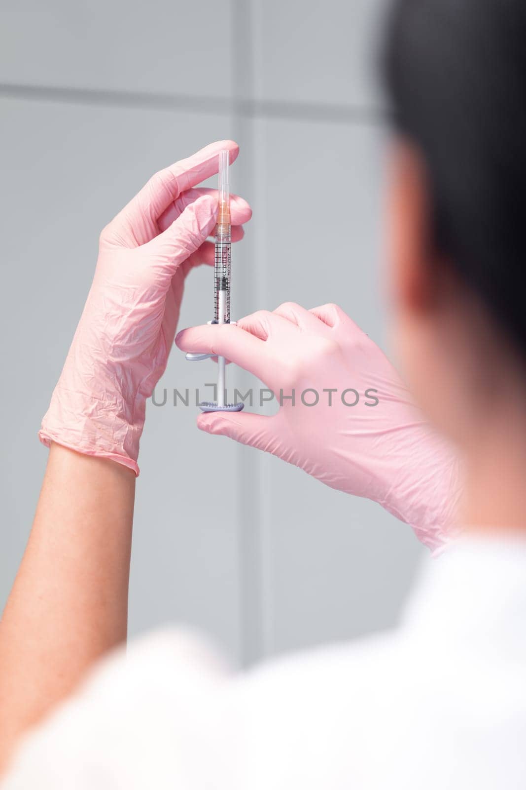 Woman cosmetologist checking for air bubbles in syringe. Back view of anonymous doctor in pink gloves holding injection preparing for beauty treatment in clinic