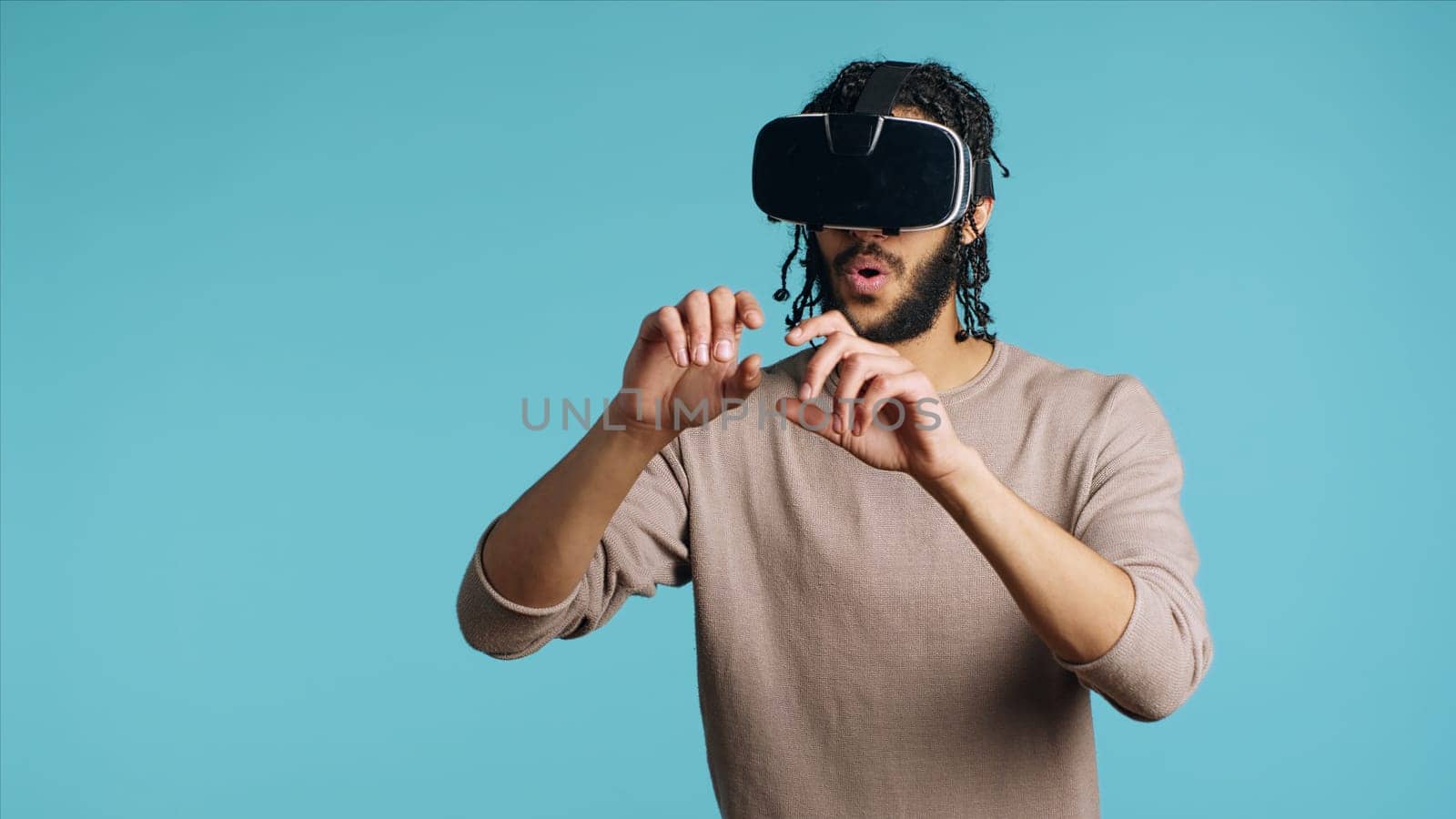 Middle Eastern man wearing virtual reality headset, doing swiping gestures. BIPOC person using high tech futuristic modern VR gadget, isolated over blue studio background, camera A