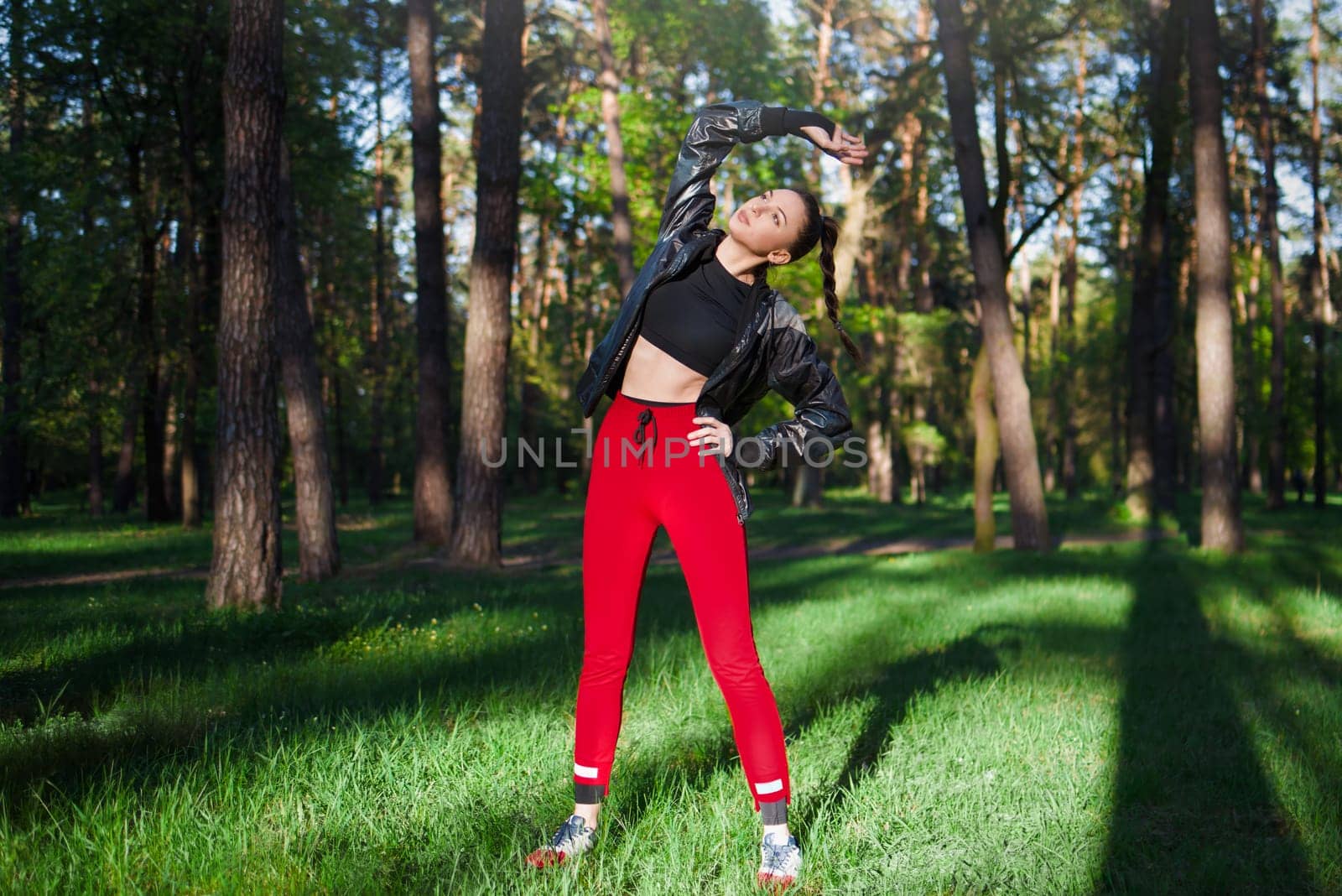 Active woman in red sportswear stretches before a run in a sunny green park