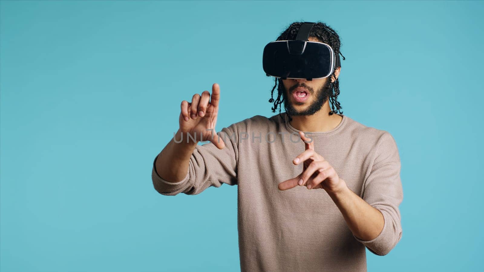 Tech enthusiast wearing virtual reality goggles, doing swiping gestures. BIPOC man using high tech futuristic modern VR glasses, making tactile touching gesturing, blue studio background
