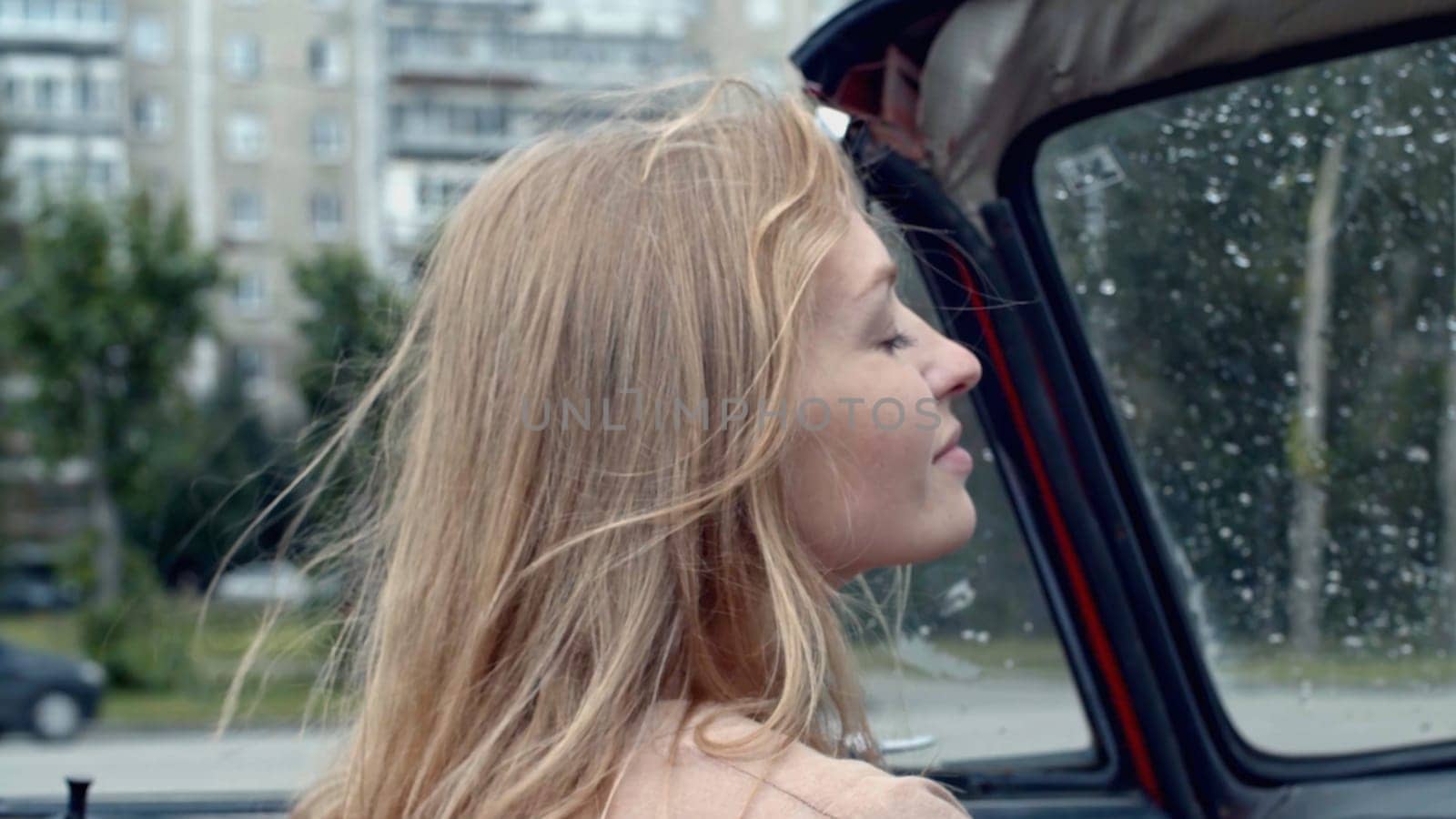 Young woman on passenger seat in a cabriolet car. Stock. Rain drops on car windshield and summer city on the background