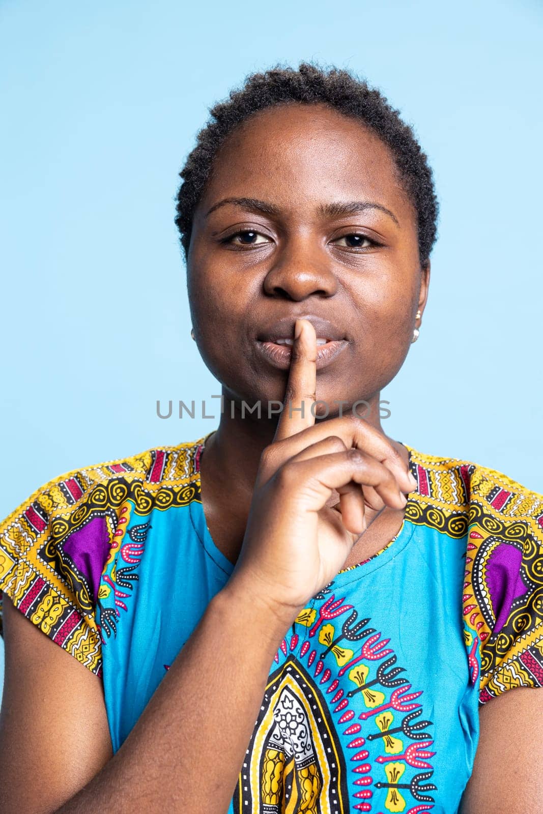 African american girl doing silence privacy symbol in studio, showing mute gesture to keep secret and be private. Young silent person doing hush sign over blue background, confidential information.