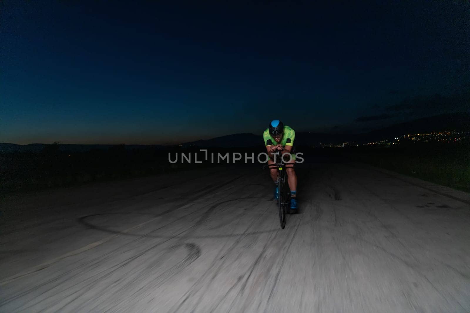 A triathlete rides his bike in the darkness of night, pushing himself to prepare for a marathon. The contrast between the darkness and the light of his bike creates a sense of drama and highlights the athlete's determination and perseverance