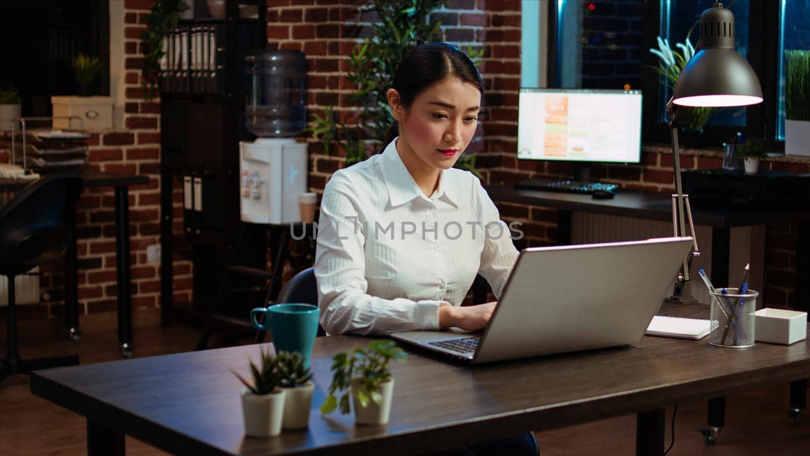 Smiling woman sitting down at office desk, typing on laptop keyboard, starting work with mug full of coffee, solving tasks late at night. Cheerful employee imputing data on computer