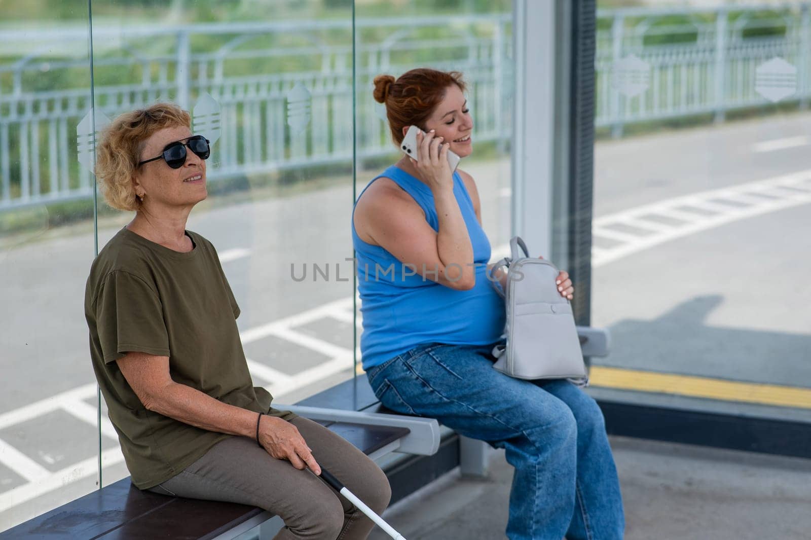 An elderly blind woman and a pregnant woman are sitting at a bus stop and waiting for the bus. by mrwed54