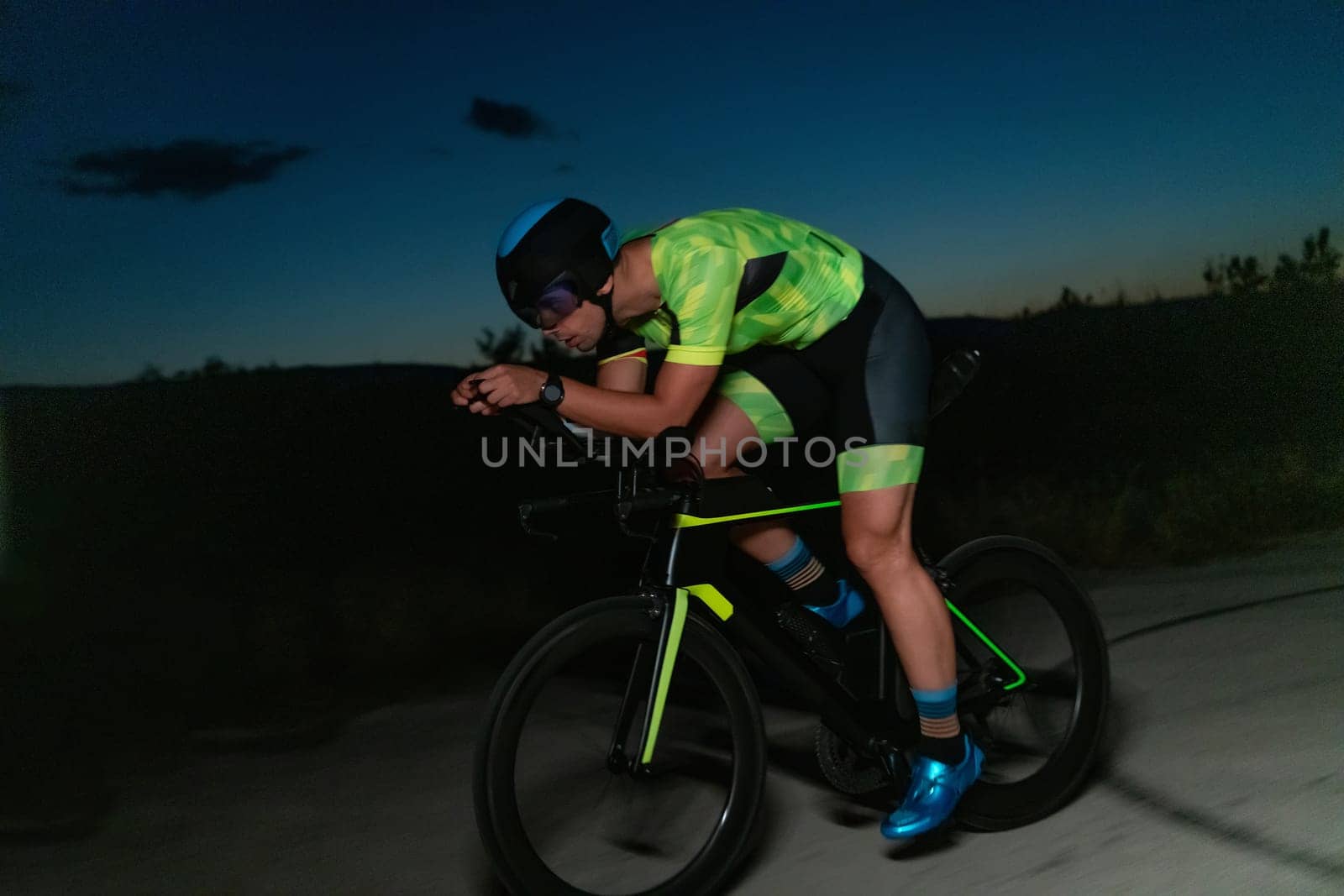 A triathlete rides his bike in the darkness of night, pushing himself to prepare for a marathon. The contrast between the darkness and the light of his bike creates a sense of drama and highlights the athlete's determination and perseverance