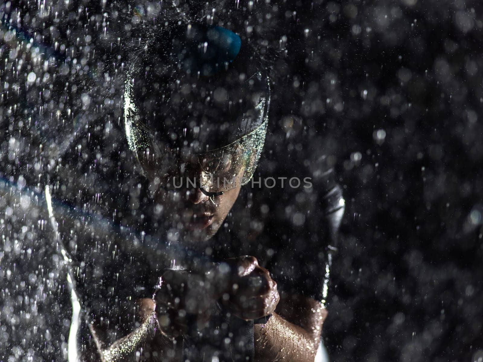 A triathlete braving the rain as he cycles through the night, preparing himself for the upcoming marathon. The blurred raindrops in the foreground and the dark, moody atmosphere in the background add to the sense of determination and grit shown by the athlete