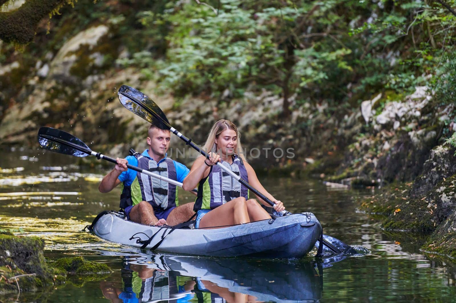 A young couple enjoying an idyllic kayak ride in the middle of a beautiful river surrounded by forest greenery by dotshock