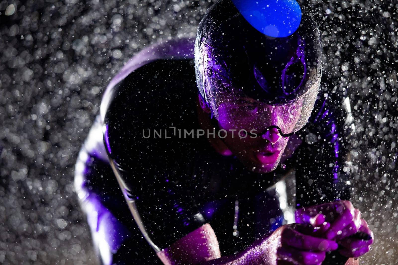 A triathlete braving the rain as he cycles through the night, preparing himself for the upcoming marathon. The blurred raindrops in the foreground and the dark, moody atmosphere in the background add to the sense of determination and grit shown by the athlete