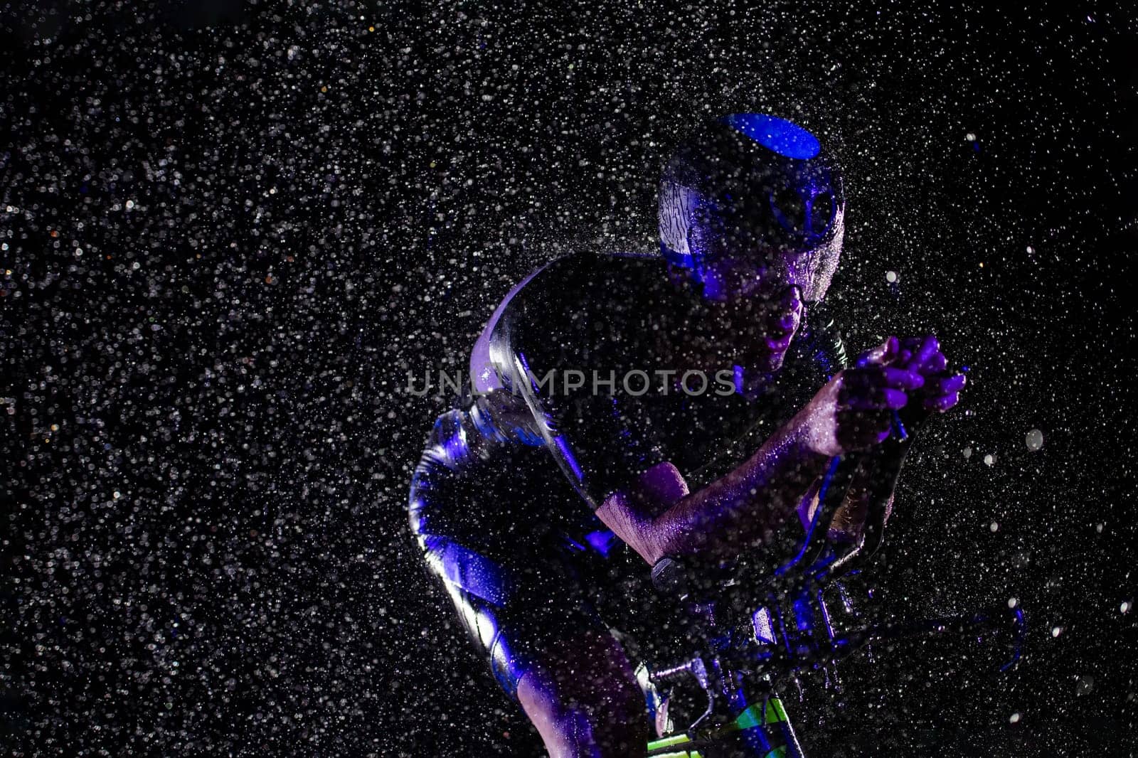 A triathlete braving the rain as he cycles through the night, preparing himself for the upcoming marathon. The blurred raindrops in the foreground and the dark, moody atmosphere in the background add to the sense of determination and grit shown by the athlete