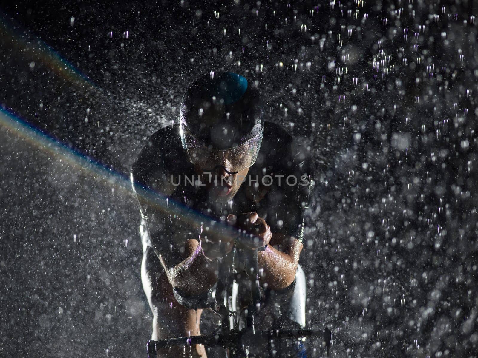 A triathlete braving the rain as he cycles through the night, preparing himself for the upcoming marathon. The blurred raindrops in the foreground and the dark, moody atmosphere in the background add to the sense of determination and grit shown by the athlete