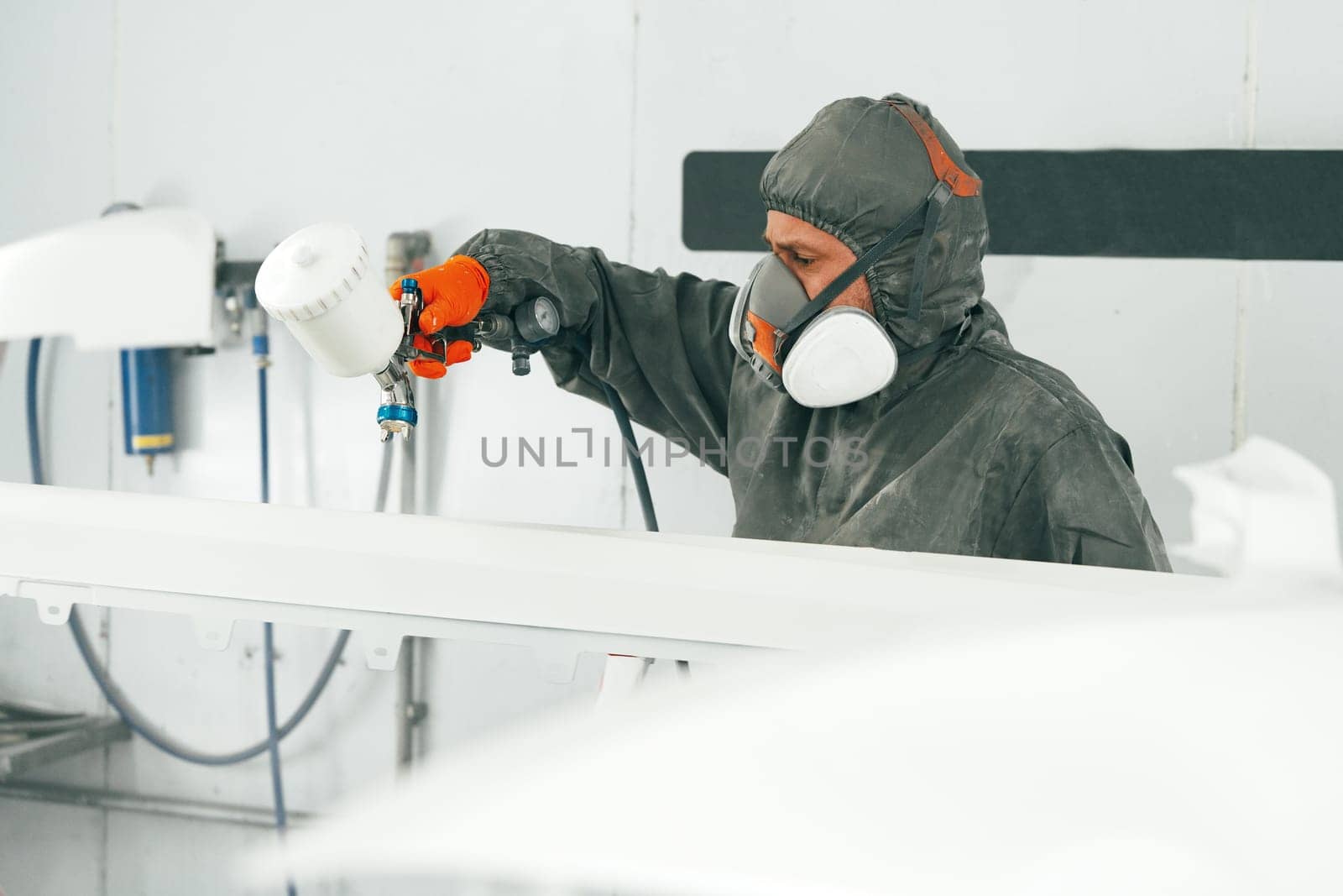 Male painter in respiratory mask and protective suit painting car with spray gun in car service close up