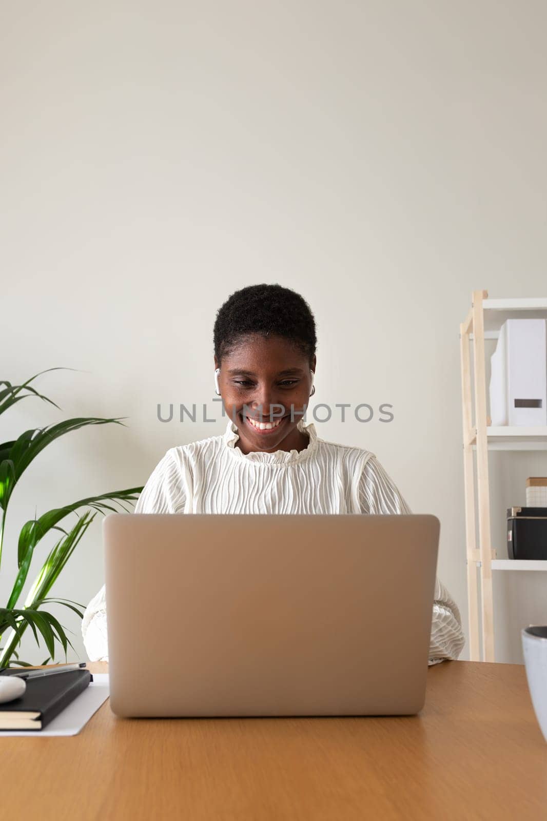 Vertical portrait of African american woman working at home office. Happy Black female entrepreneur using laptop and wireless earphones. Working from home concept.