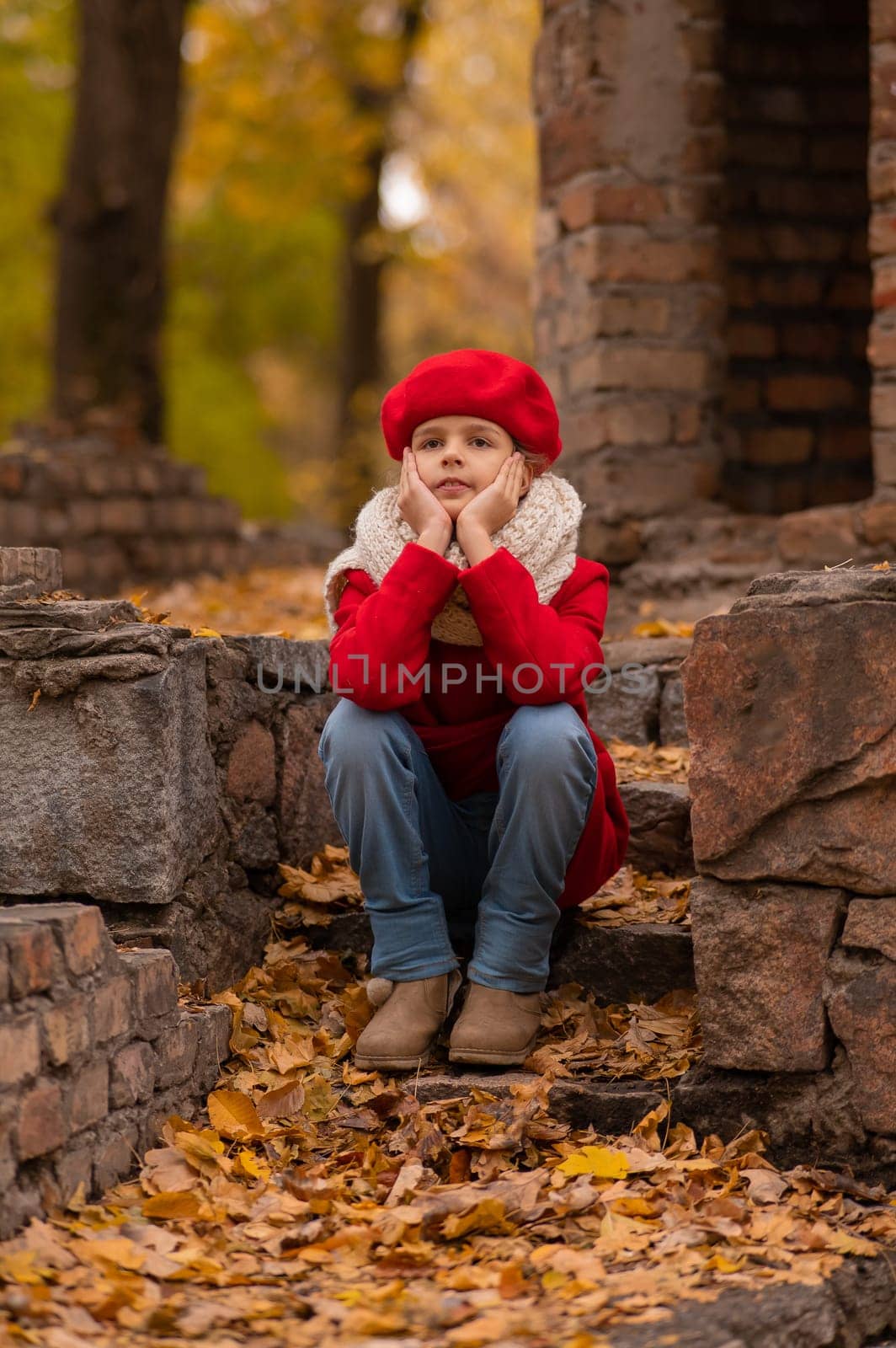 Sad caucasian girl in a red coat and beret sits on a brick wall on a walk in autumn