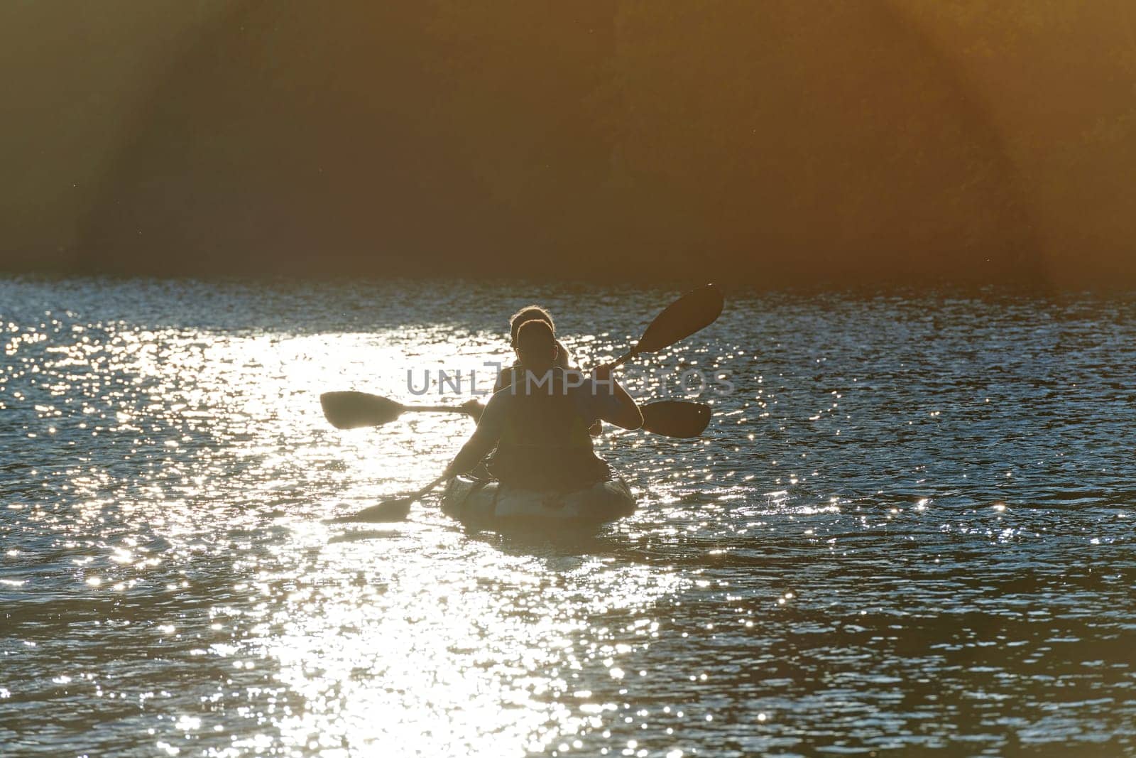 A young couple enjoying an idyllic kayak ride in the middle of a beautiful river surrounded by forest greenery in sunset time.