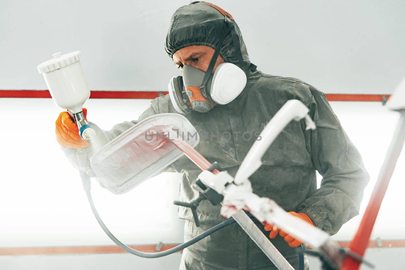 Male painter in respiratory mask and protective suit painting car with spray gun in car service close up